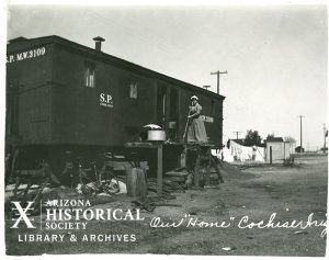 Woman holding a broom standing on platform entrance to boxcar home. Text written on image reads "Our "home" Cochise, Ariz"